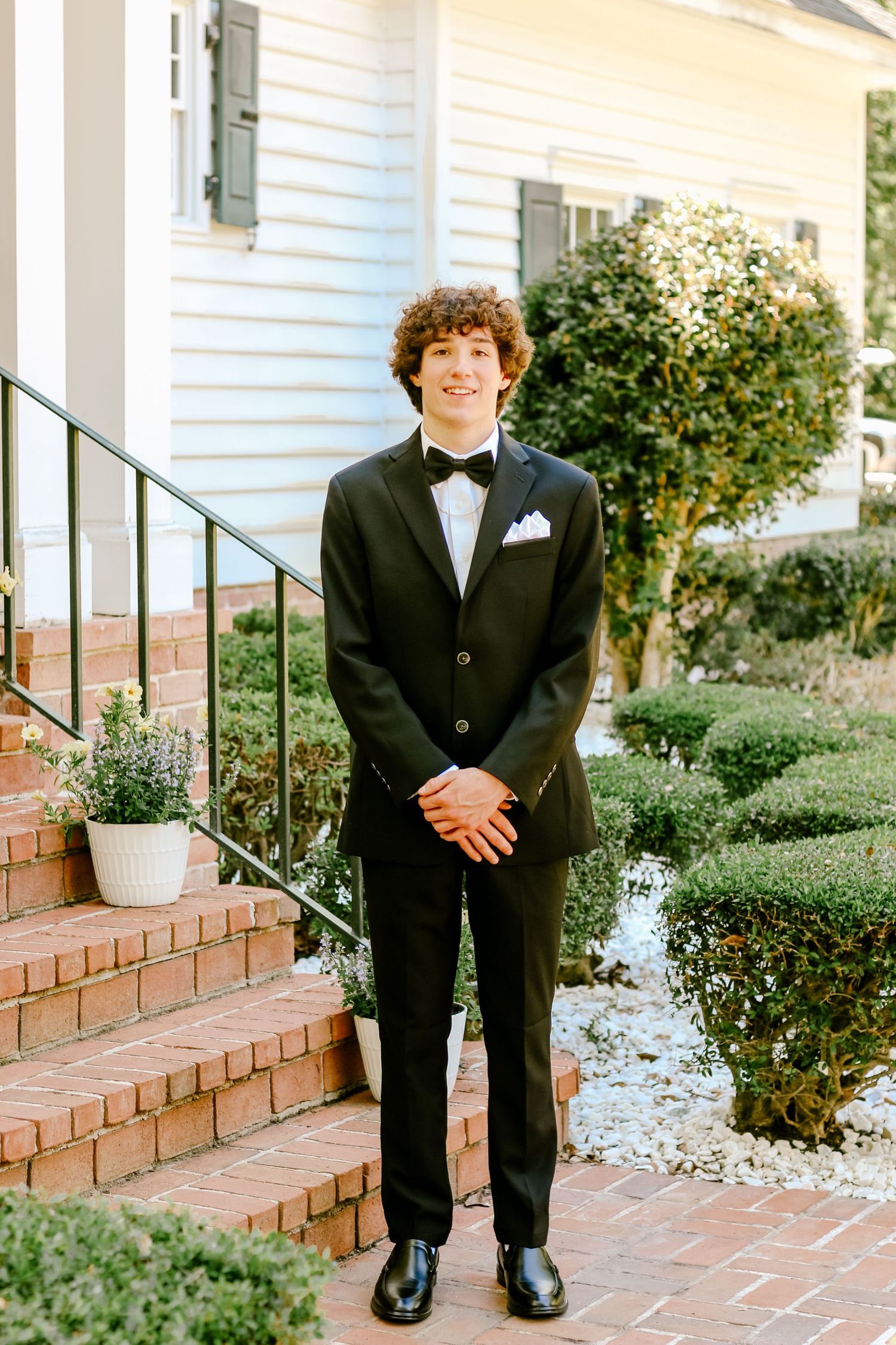 Person in classic black tuxedo poses on brick steps of white colonial style home.