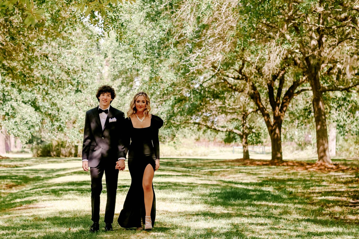 A formal couple walking together on a tree-lined path in elegant attire.