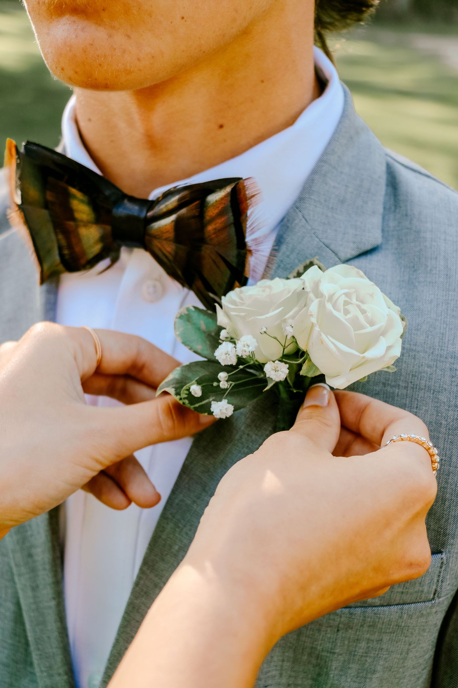 Hands pinning a classic white rose boutonniere onto a grey suit lapel.