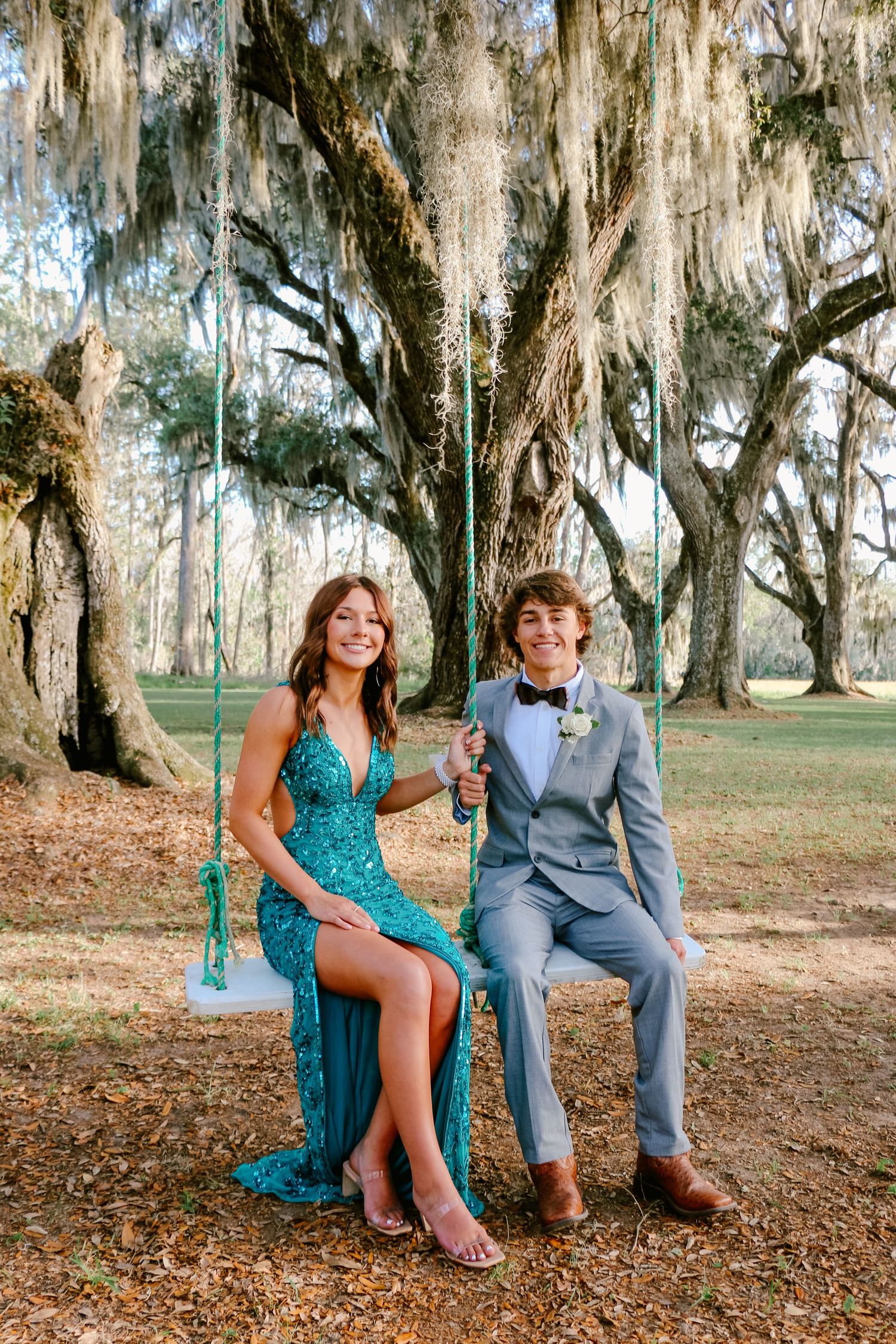 A couple in formal wear sits on a swing beneath moss-covered oak trees in a romantic Southern setting.