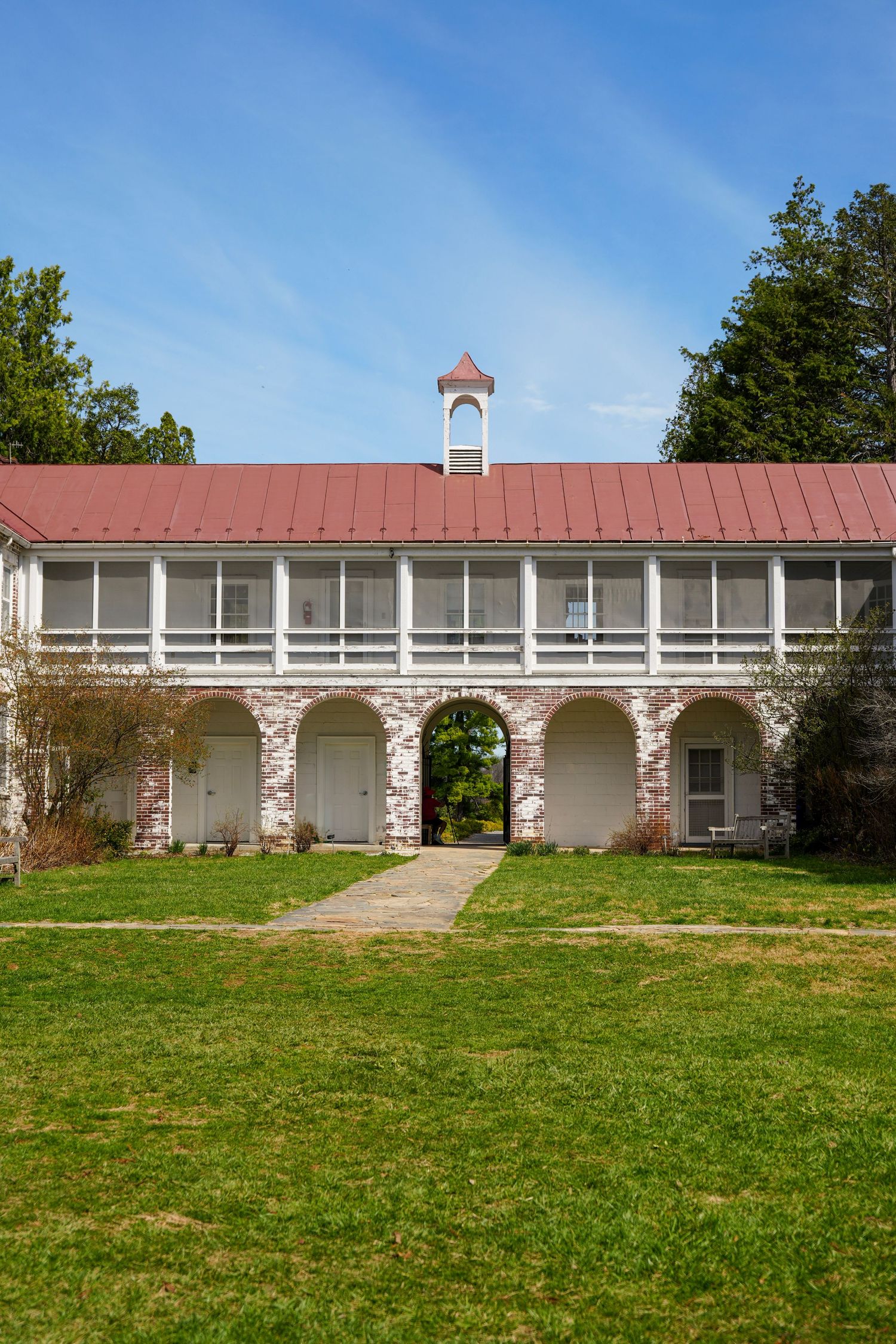 Historic two-story stone building with red metal roof, white upper balcony, arched walkway, and bell tower on sunny day.
