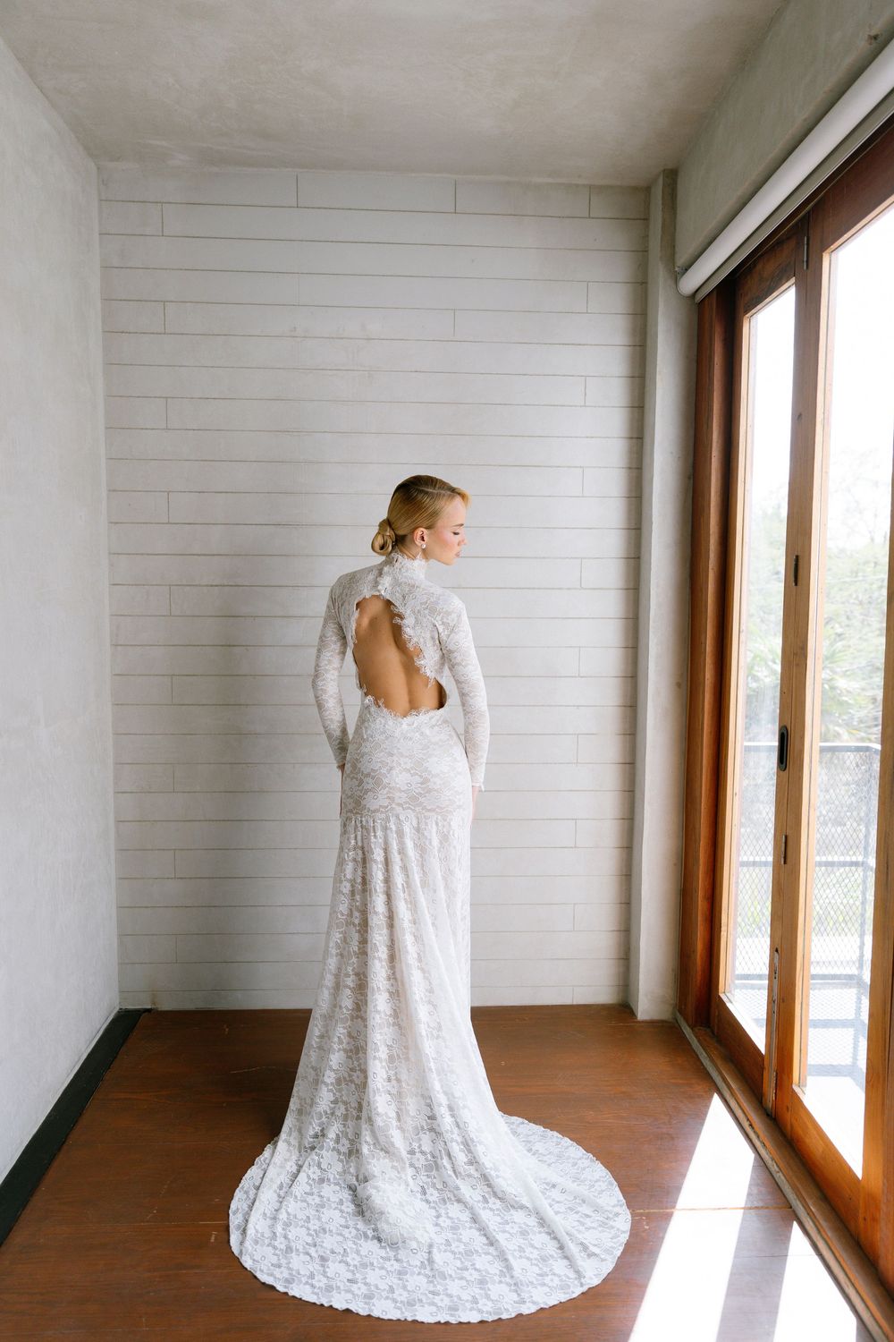 Bride in elegant long-sleeve lace wedding gown with open back and train standing by large windows.
