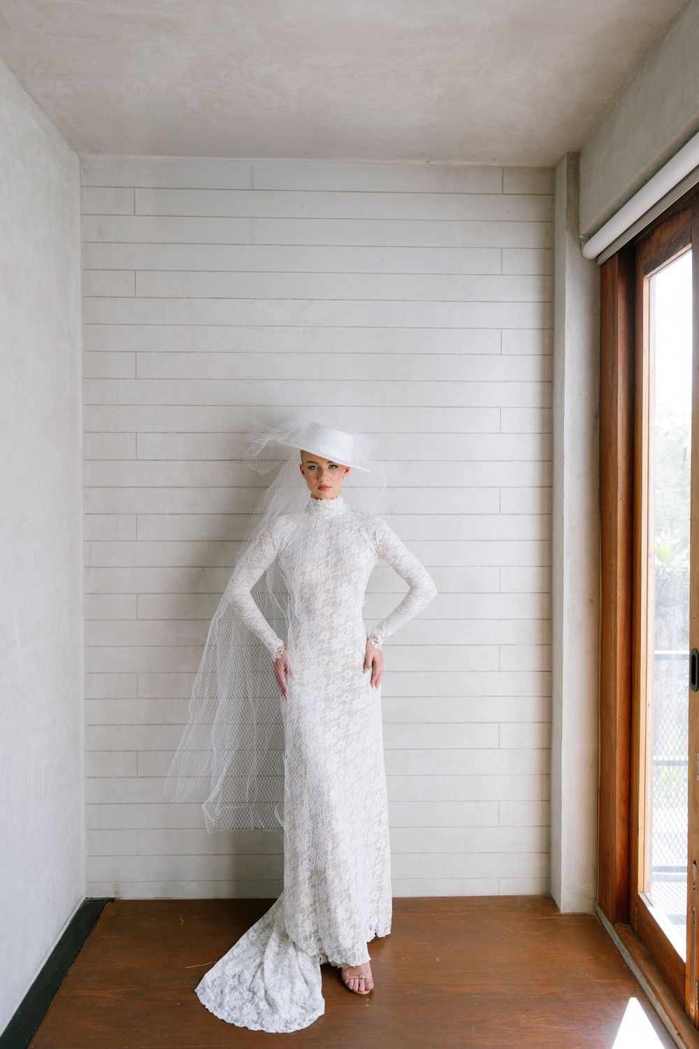 Bride wearing long-sleeve lace wedding gown with wide-brim white hat and veil indoors.