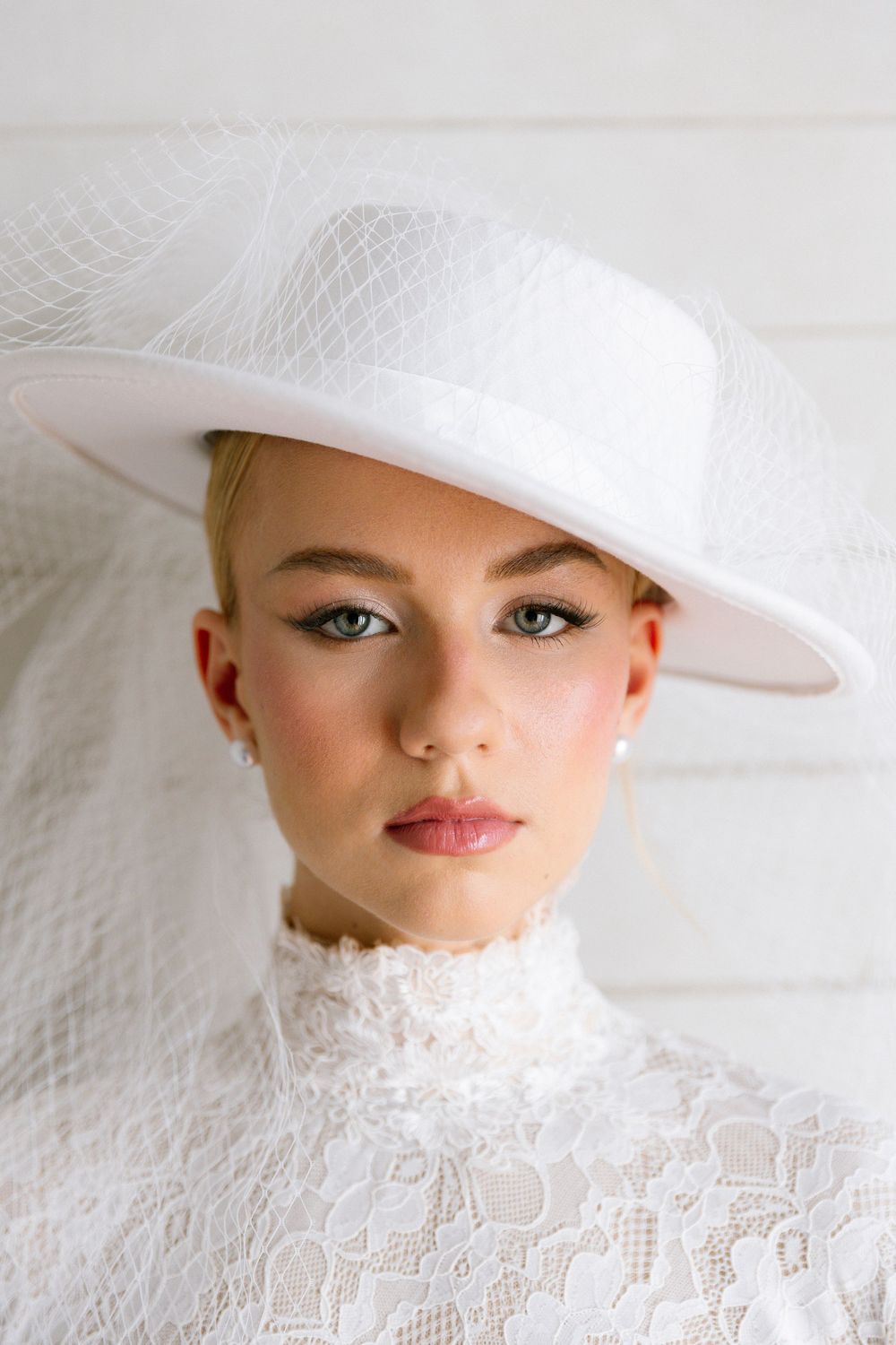 Close-up portrait of bride wearing white wide-brim hat with tulle veil and lace dress.