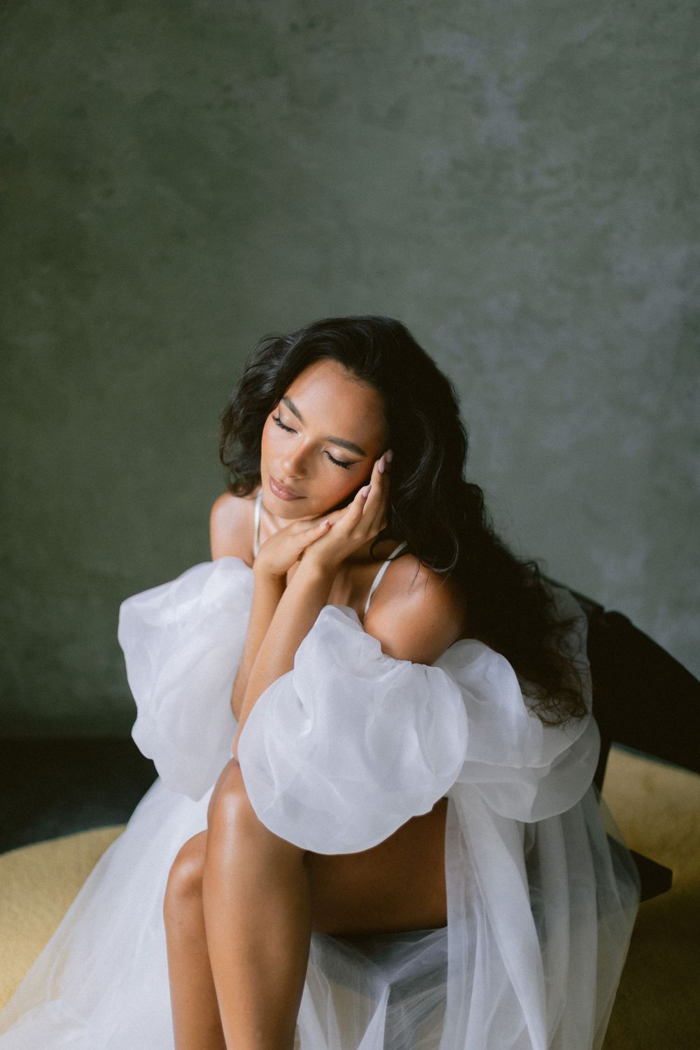 Beautiful dark-haired bride in white ruffled boudoir robe sitting peacefully with eyes closed.
