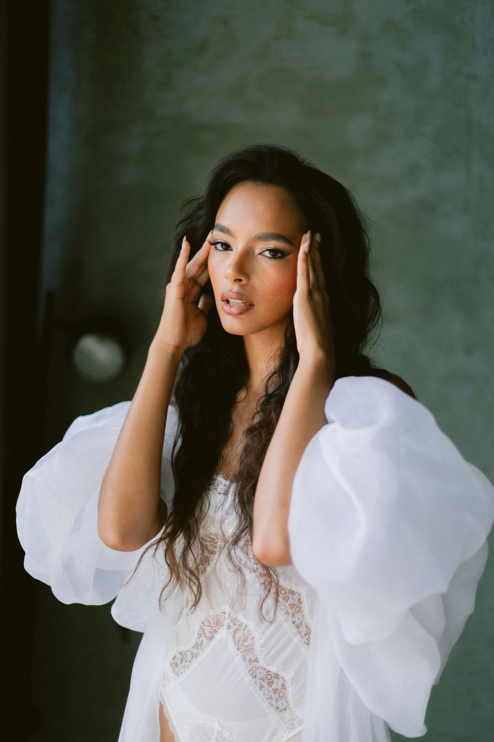 Stunning dark-haired bride in white lace robe with dramatic puff sleeves posing indoors.