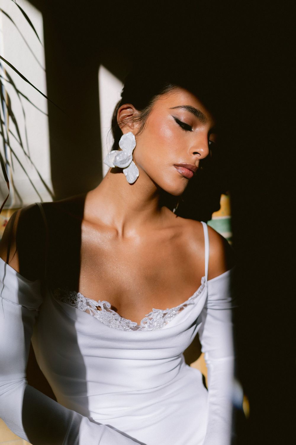 Bride in white satin slip dress with lace trim and large floral earrings, bathed in dramatic shadow and light.