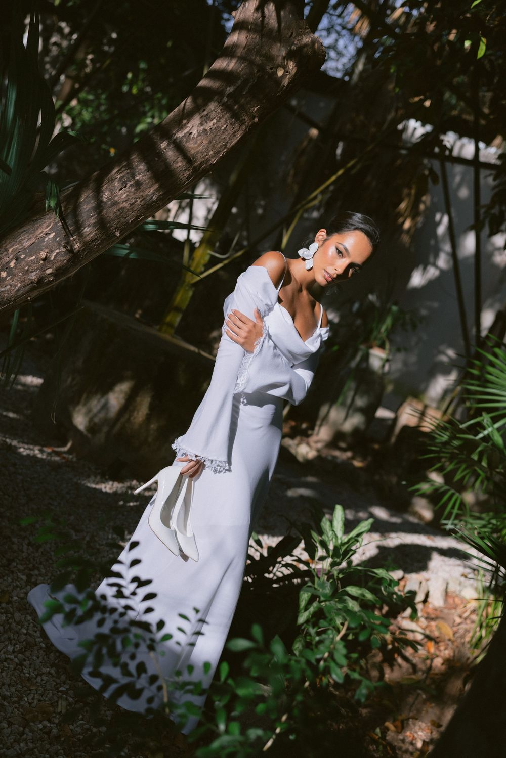 Bride in white off-shoulder fitted gown with bell sleeves posing among lush tropical garden greenery and trees.