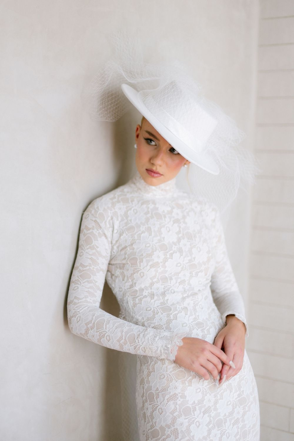 Bride in long-sleeve white lace dress wearing elegant wide-brim white fascinator hat.