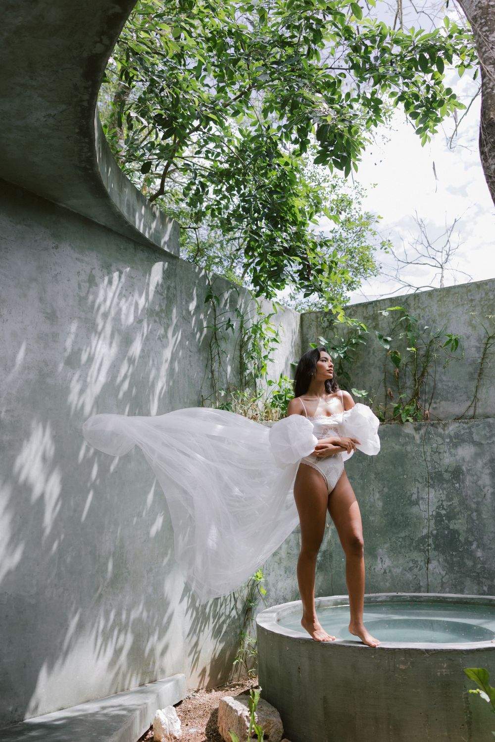 Bride in white robe standing in outdoor stone bath surrounded by lush tropical greenery.