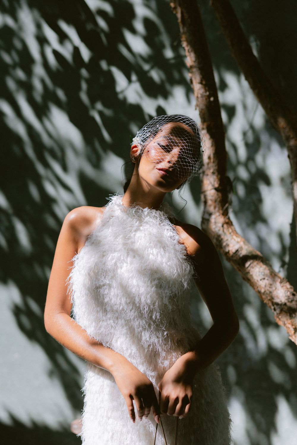 Bridal model in white feathered dress wearing a birdcage veil, leaning against a tree with leaf-shadow backdrop.