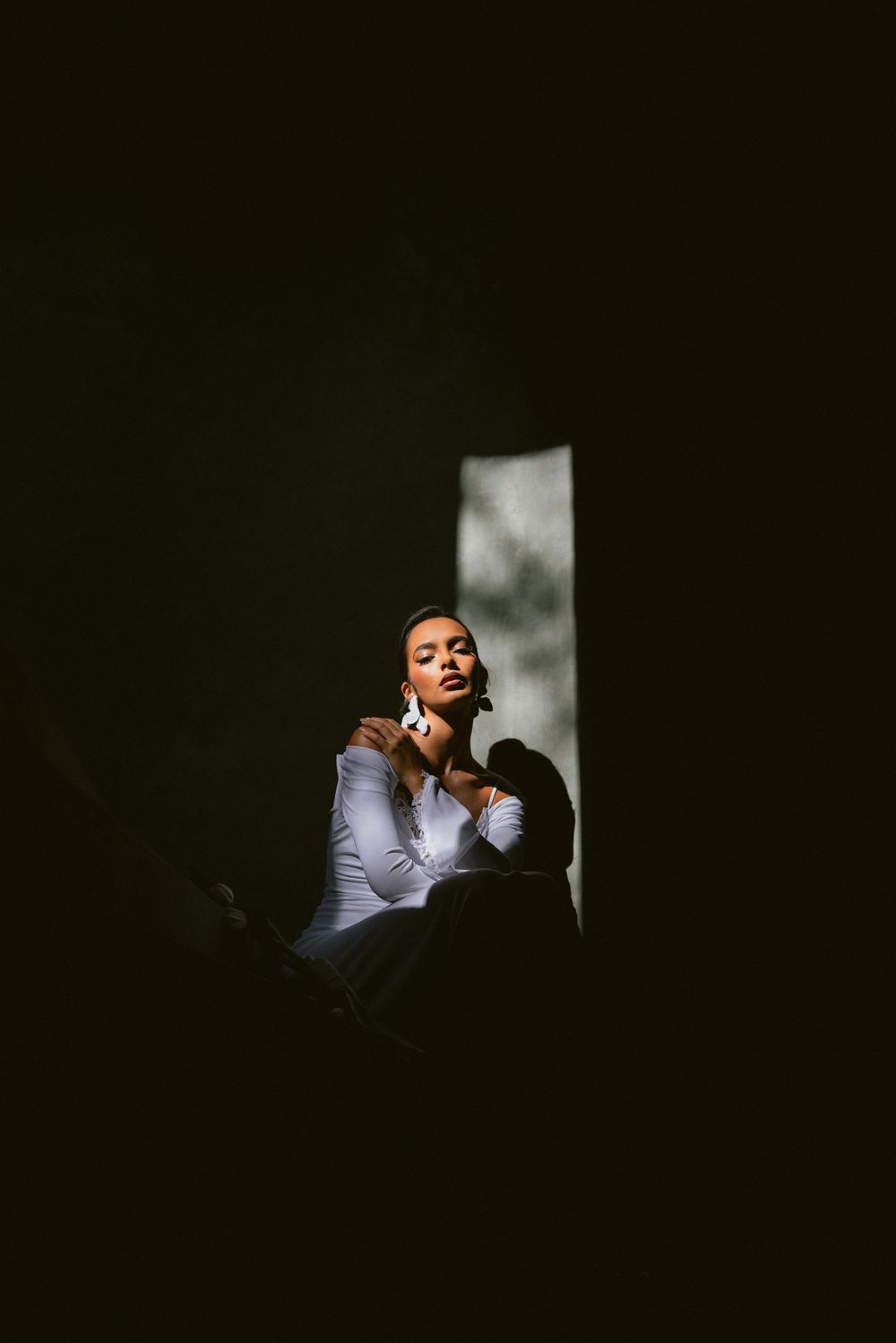Bride in white long-sleeve gown sitting in a dramatic shaft of natural light against a dark background.