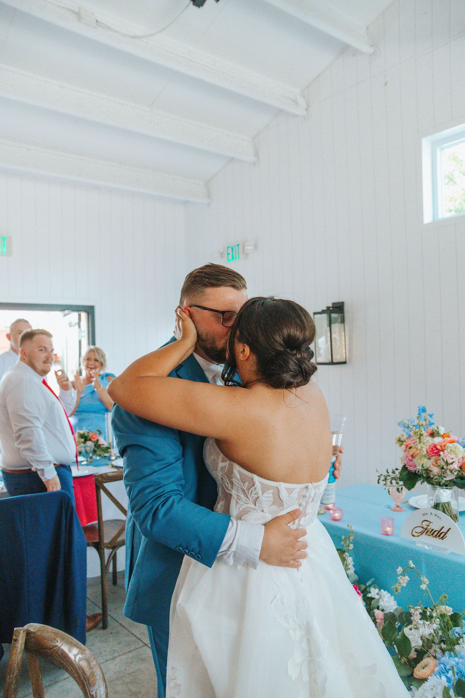 Bride and groom share a romantic first kiss at their wedding reception in a bright white venue with blue decor.