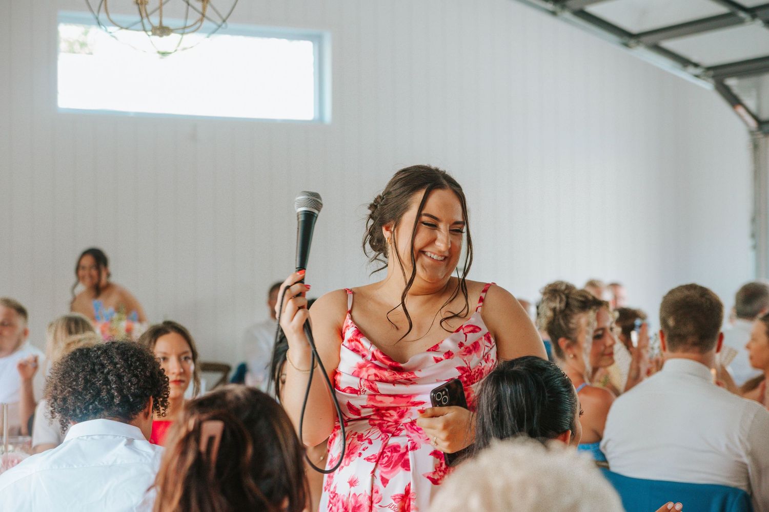 Woman in floral dress giving a speech with microphone at a bright wedding reception venue.