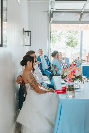 Bride and groom seated at wedding reception table with colorful floral centerpiece, guests in blue attire visible in background.