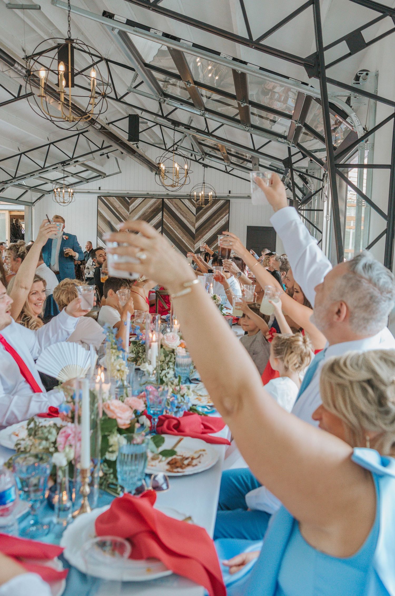 Wedding guests raising glasses in a toast at an elegant reception with floral centerpieces and chandelier lighting.