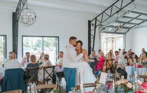 Bride and groom share first dance at rustic barn wedding reception with guests seated at decorated tables.