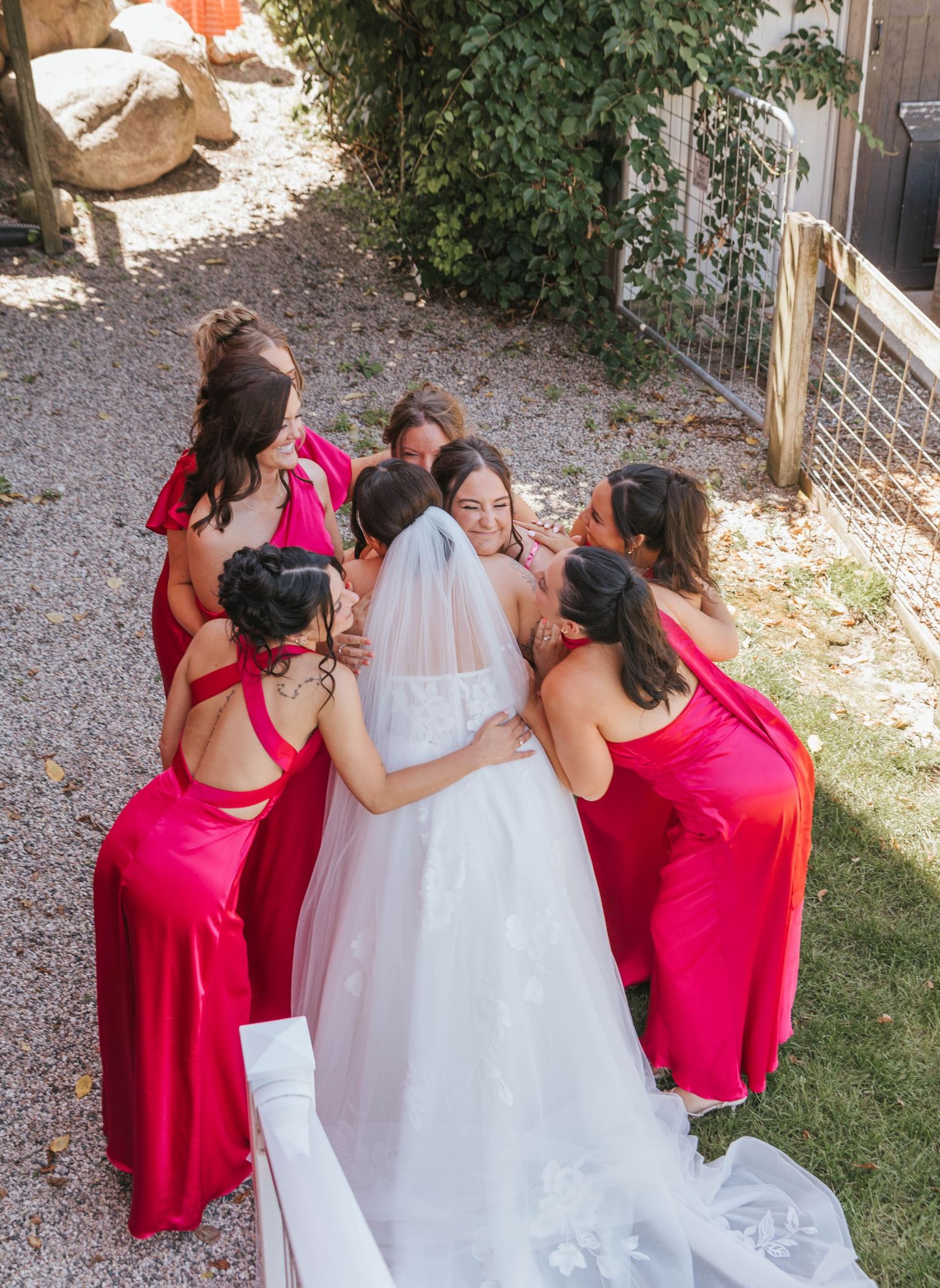 Bride in white gown and veil surrounded by bridesmaids in red dresses sharing a group hug outdoors.