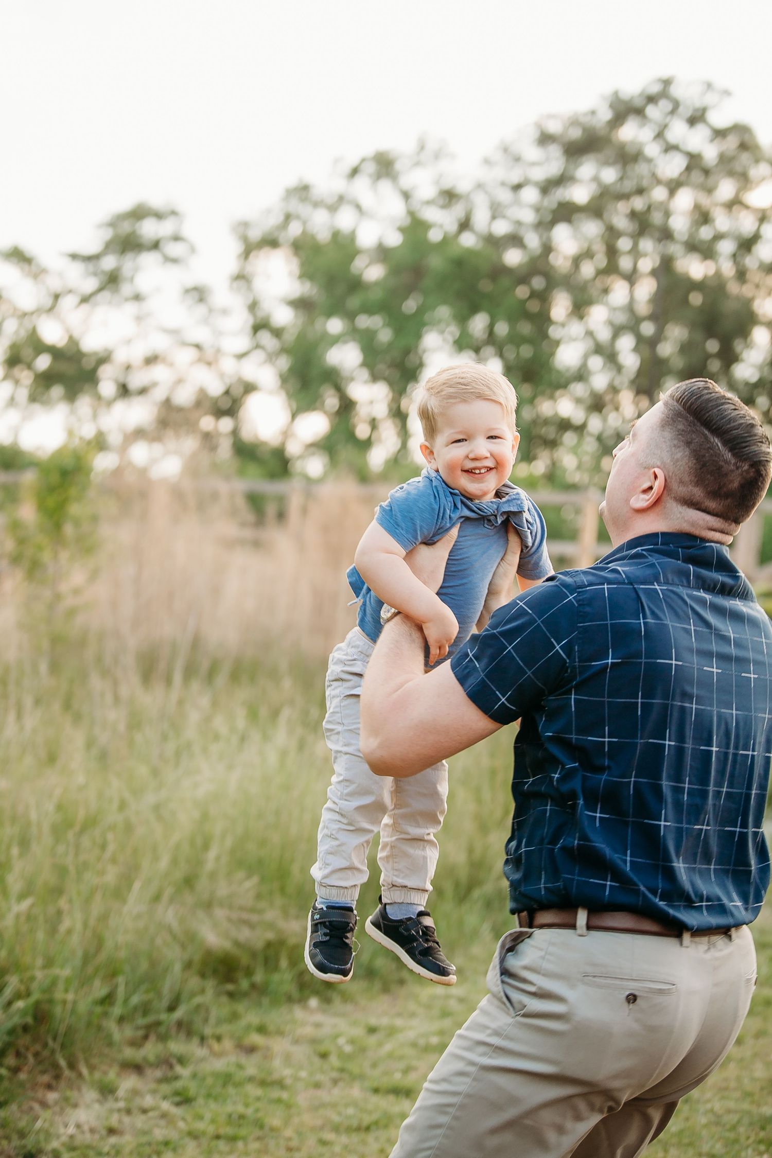 Father lifting laughing toddler boy in blue shirt outdoors in sunny green meadow.