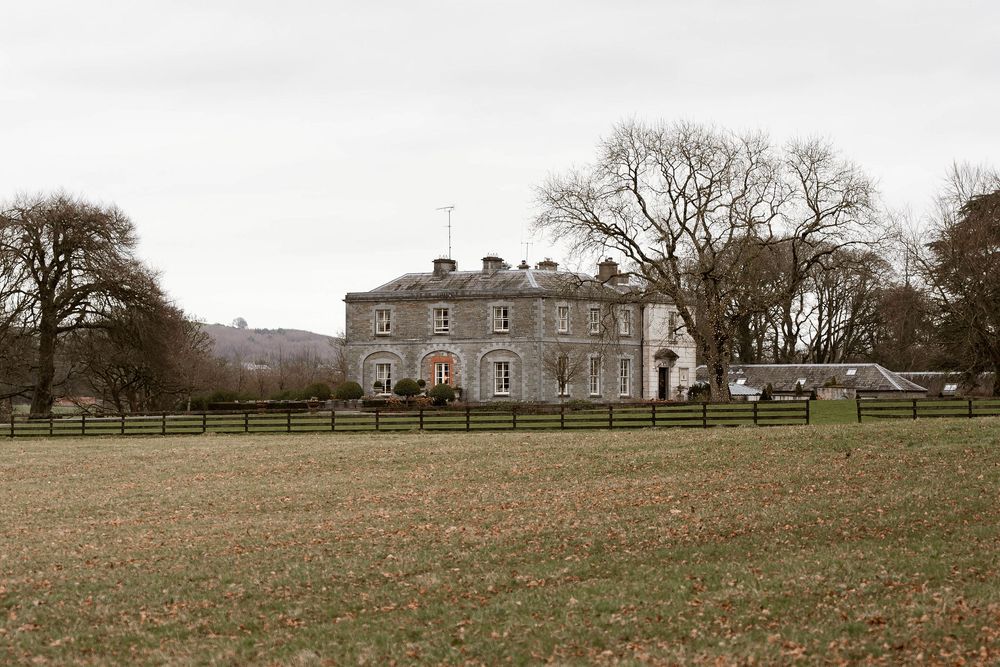 A grand stone country manor house surrounded by green fields and wooden fencing under an overcast sky.