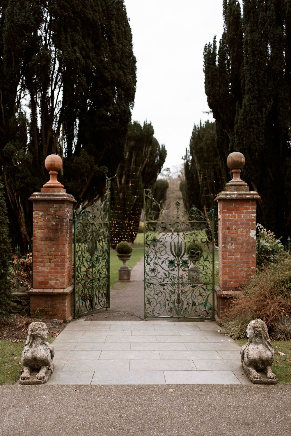 Ornate wrought iron garden gate flanked by brick pillars with stone lion statues and tall cypress trees lining a path.