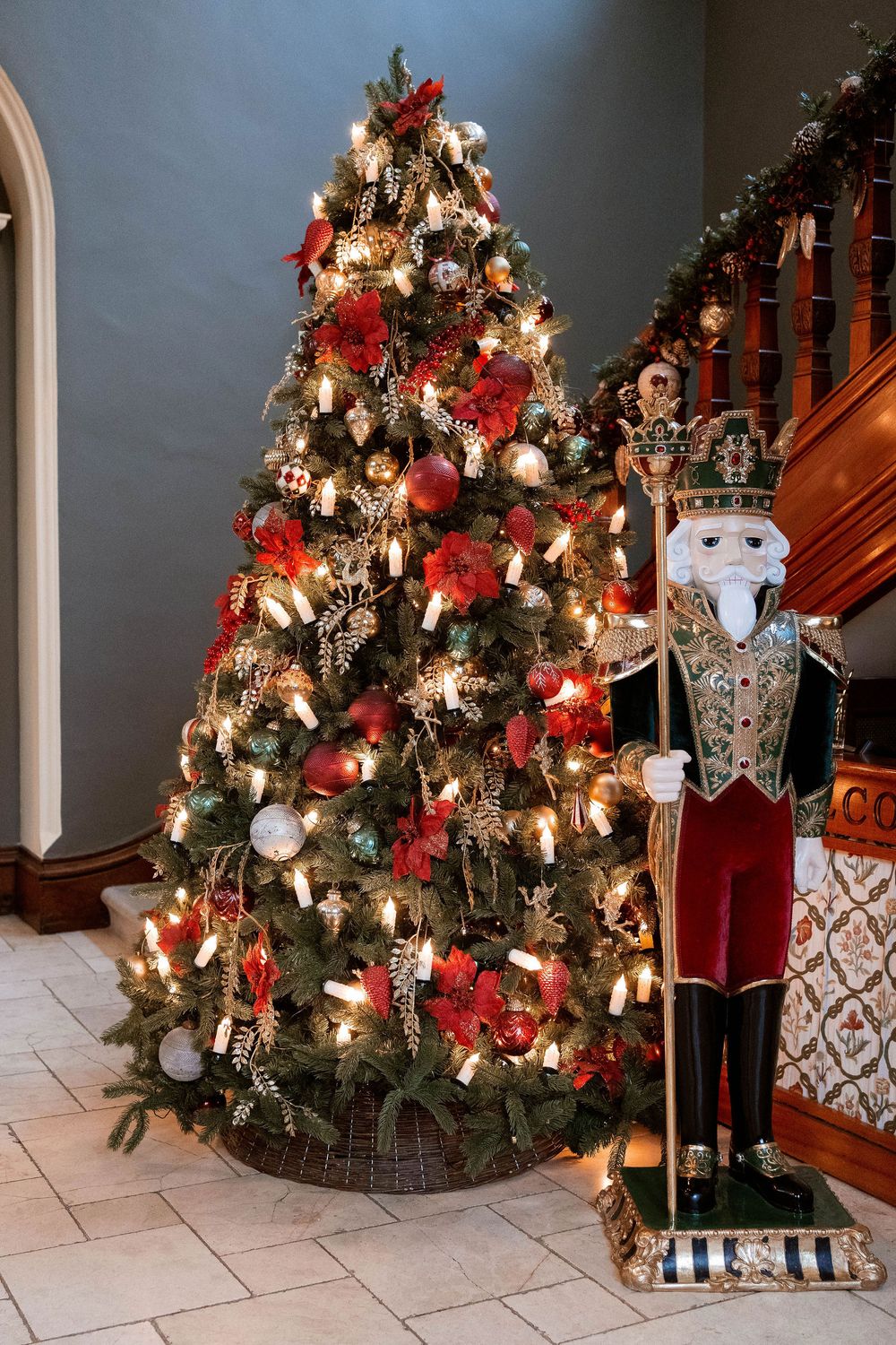 Decorated Christmas tree with red ornaments and lights beside a large nutcracker figurine near a wooden staircase.