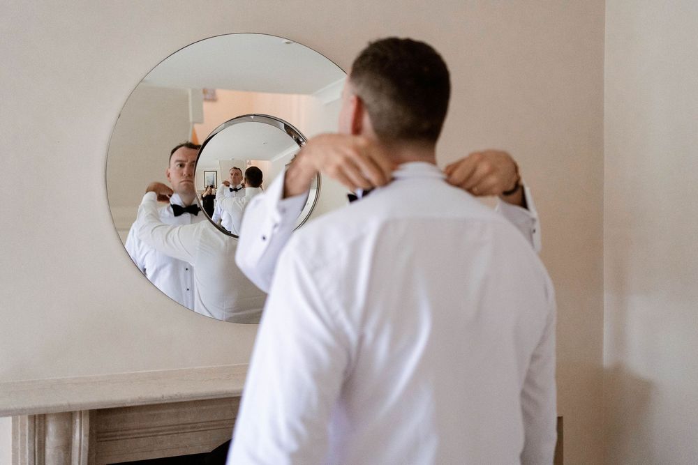 Groom in white shirt getting ready, adjusting collar while his reflection shows in a round mirror.