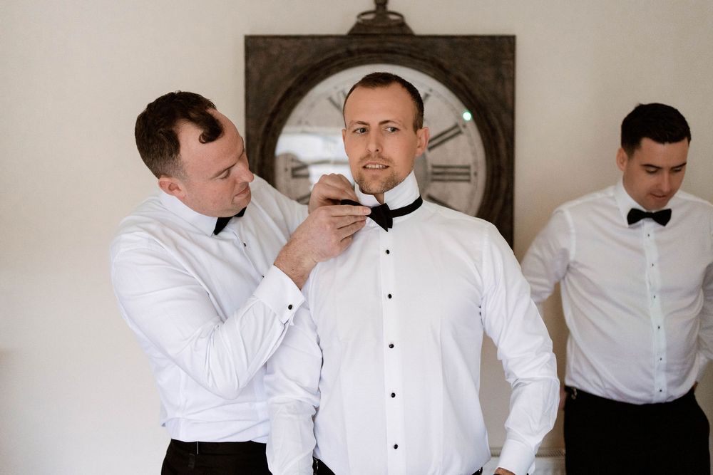 Groomsmen helping groom tie black bow tie before wedding ceremony in white dress shirts.