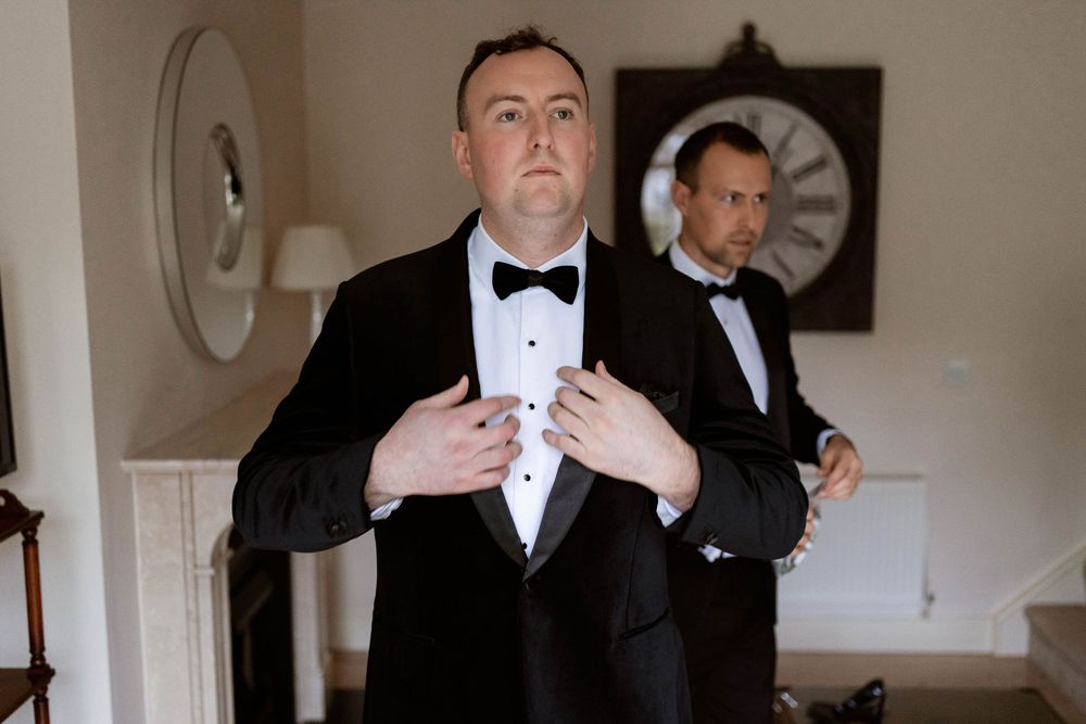 Two groomsmen in black tuxedos and bow ties prepare for a wedding ceremony indoors.