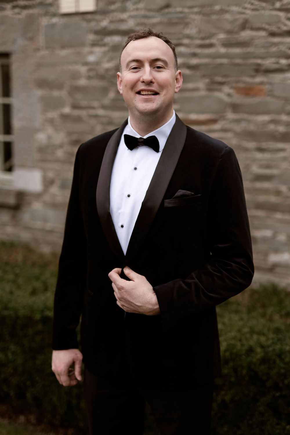 Smiling groom in black velvet tuxedo with bow tie poses outdoors against stone wall backdrop.