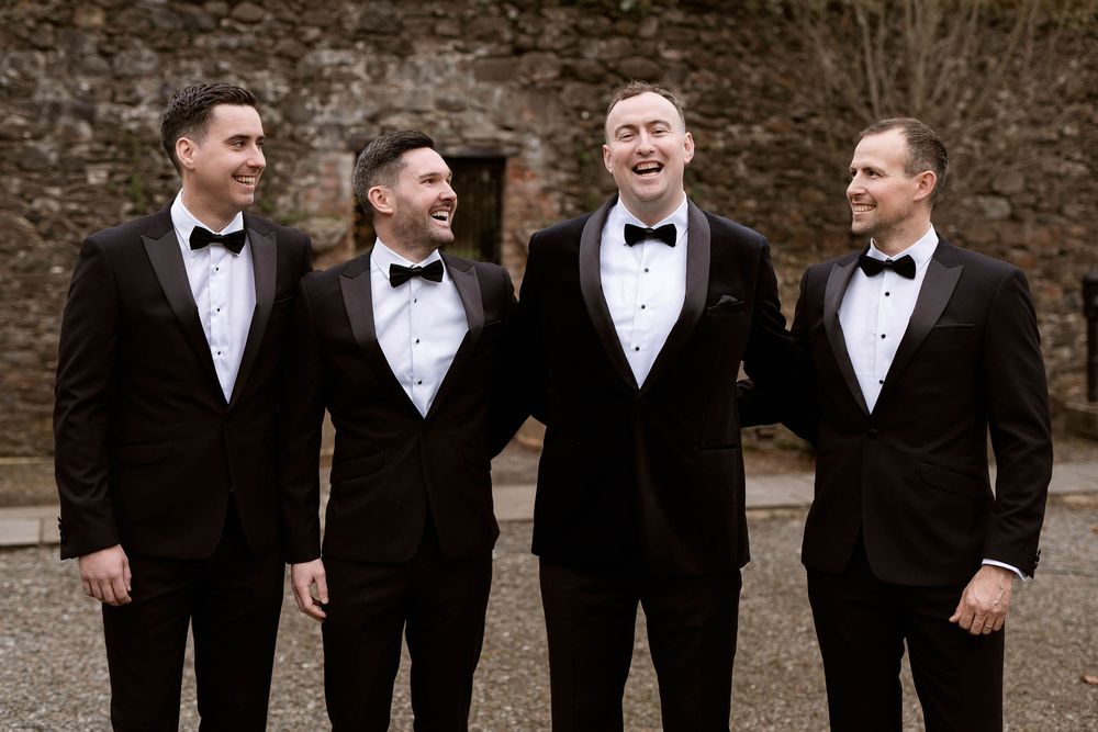 Four groomsmen in black tuxedos and bow ties laughing together outdoors at a wedding celebration.
