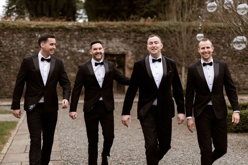 Four groomsmen in black tuxedos with bow ties walking and laughing together at a wedding venue.
