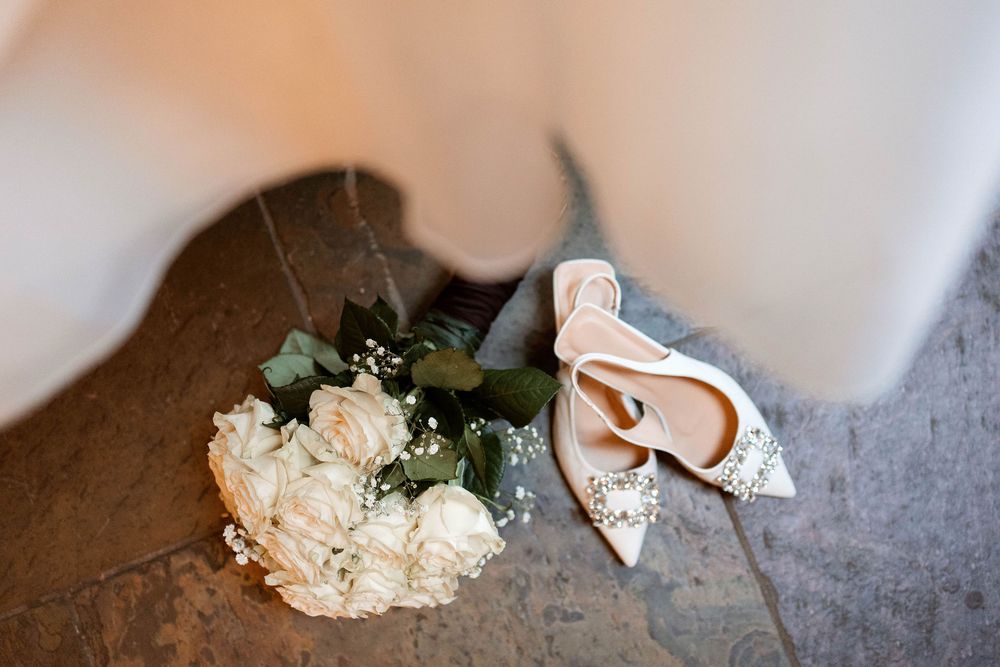 Bridal bouquet of white roses and wedding heels placed on stone floor beside flowing dress.