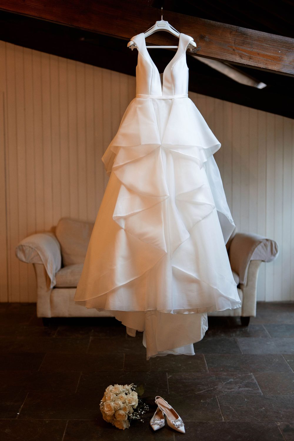 White layered ballgown wedding dress hanging on hanger with bridal bouquet and heels below.