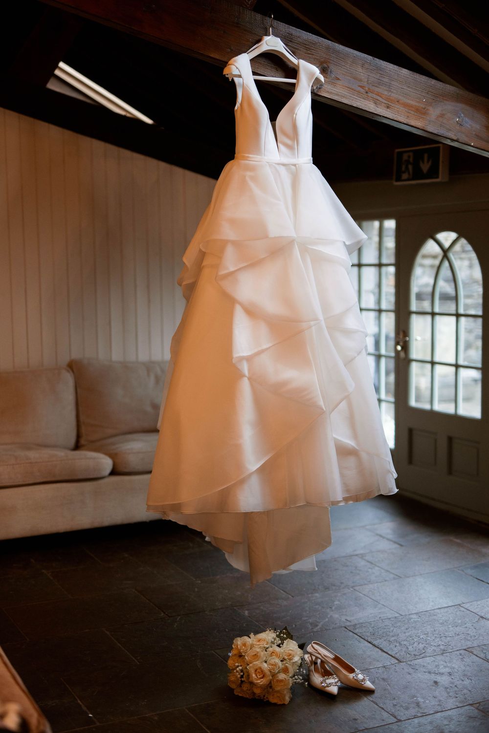 Elegant white layered wedding gown hanging in rustic venue with bridal bouquet and shoes below.