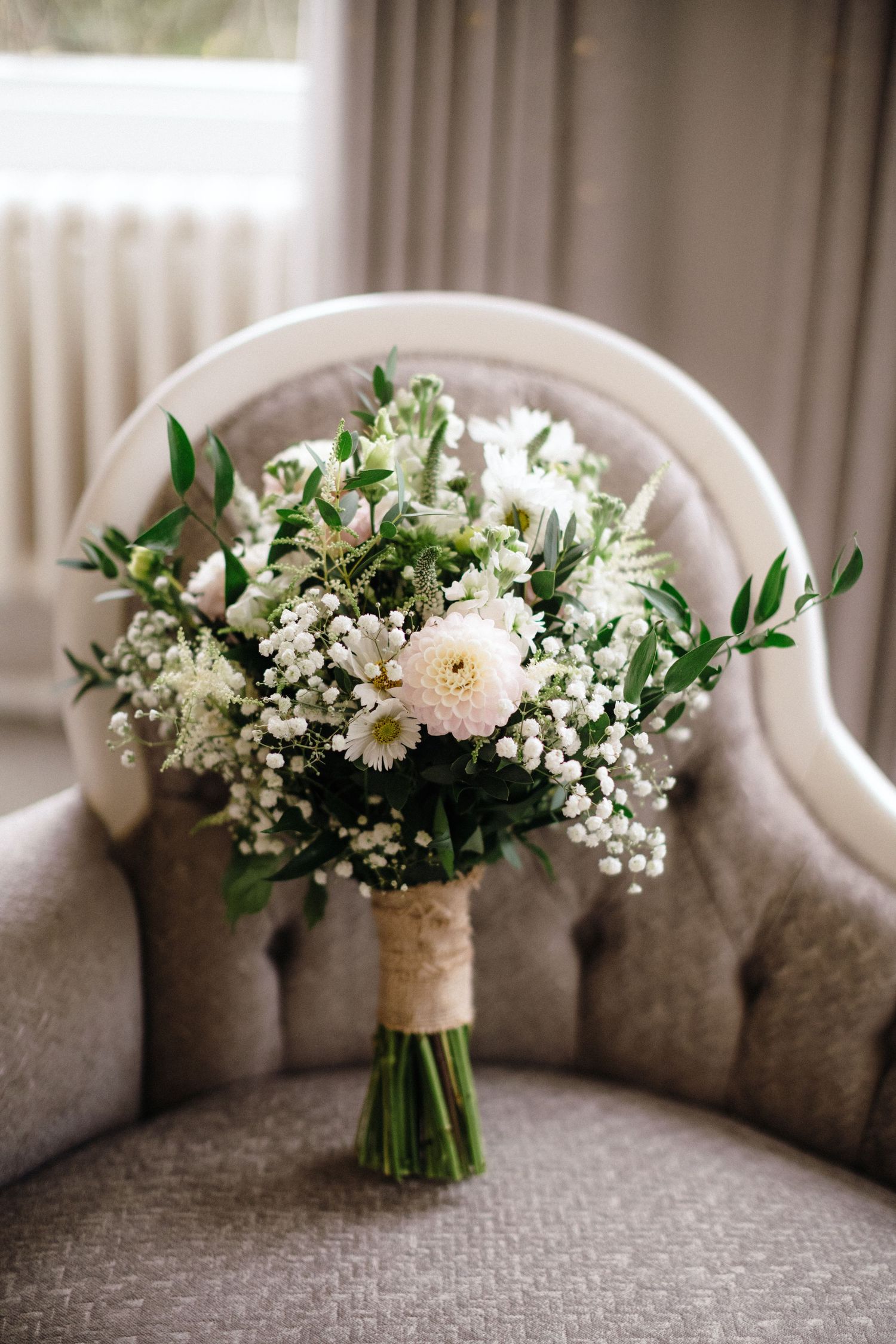 Elegant bridal bouquet of white flowers and greenery wrapped in twine, resting on a grey velvet chair.