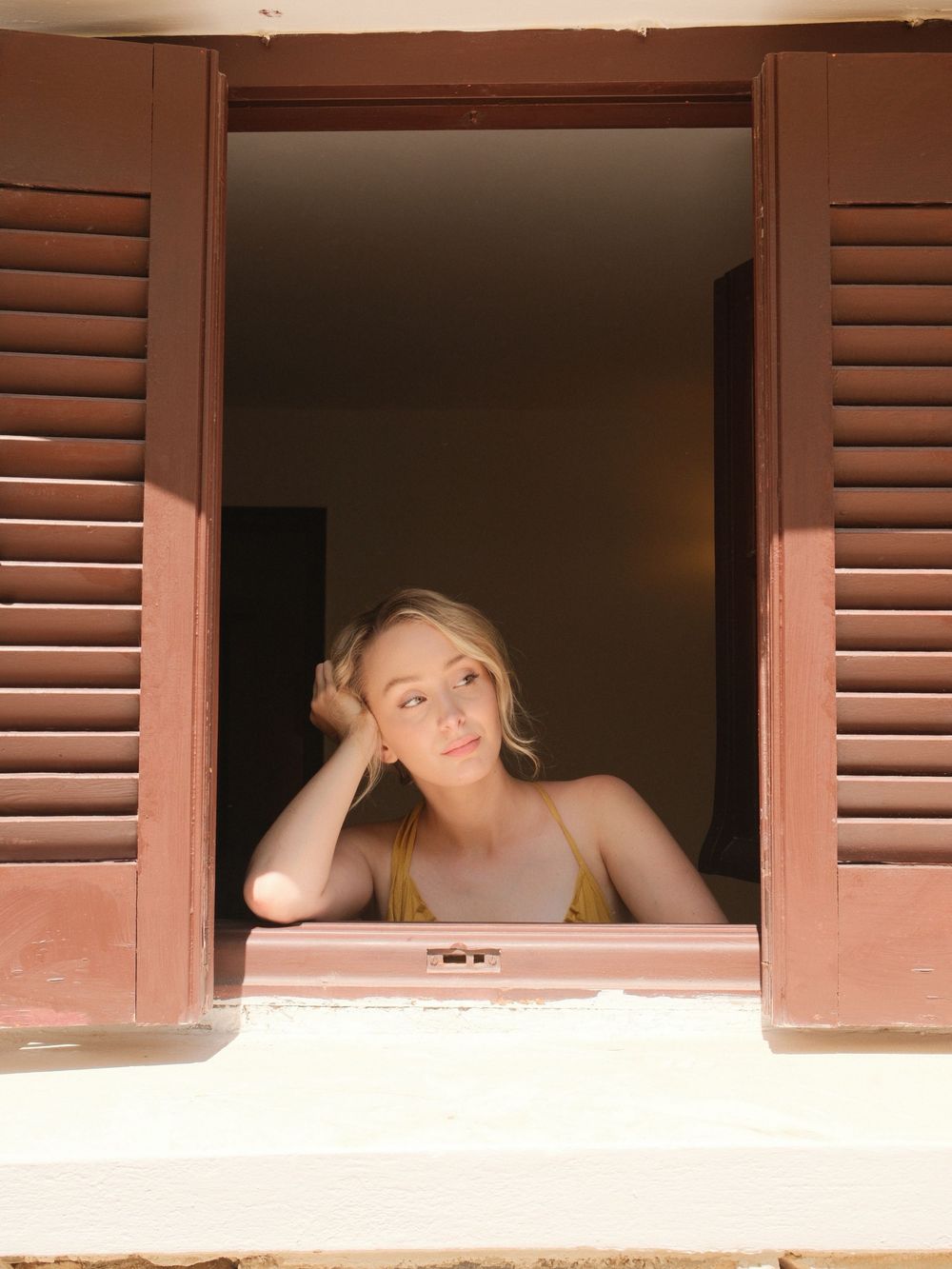 Young blonde woman resting her head on her hand, gazing out of a rustic wooden window with brown shutters.