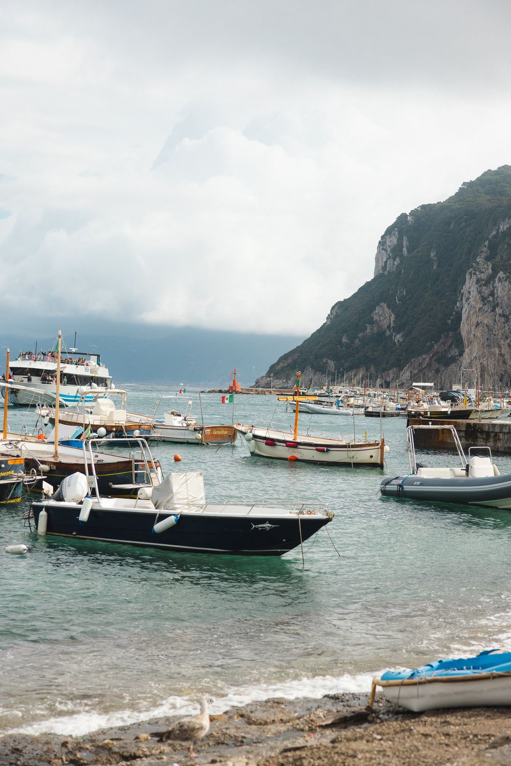Colorful boats moored in harbor at Isle of Capri with dramatic rocky cliffs under cloudy sky.