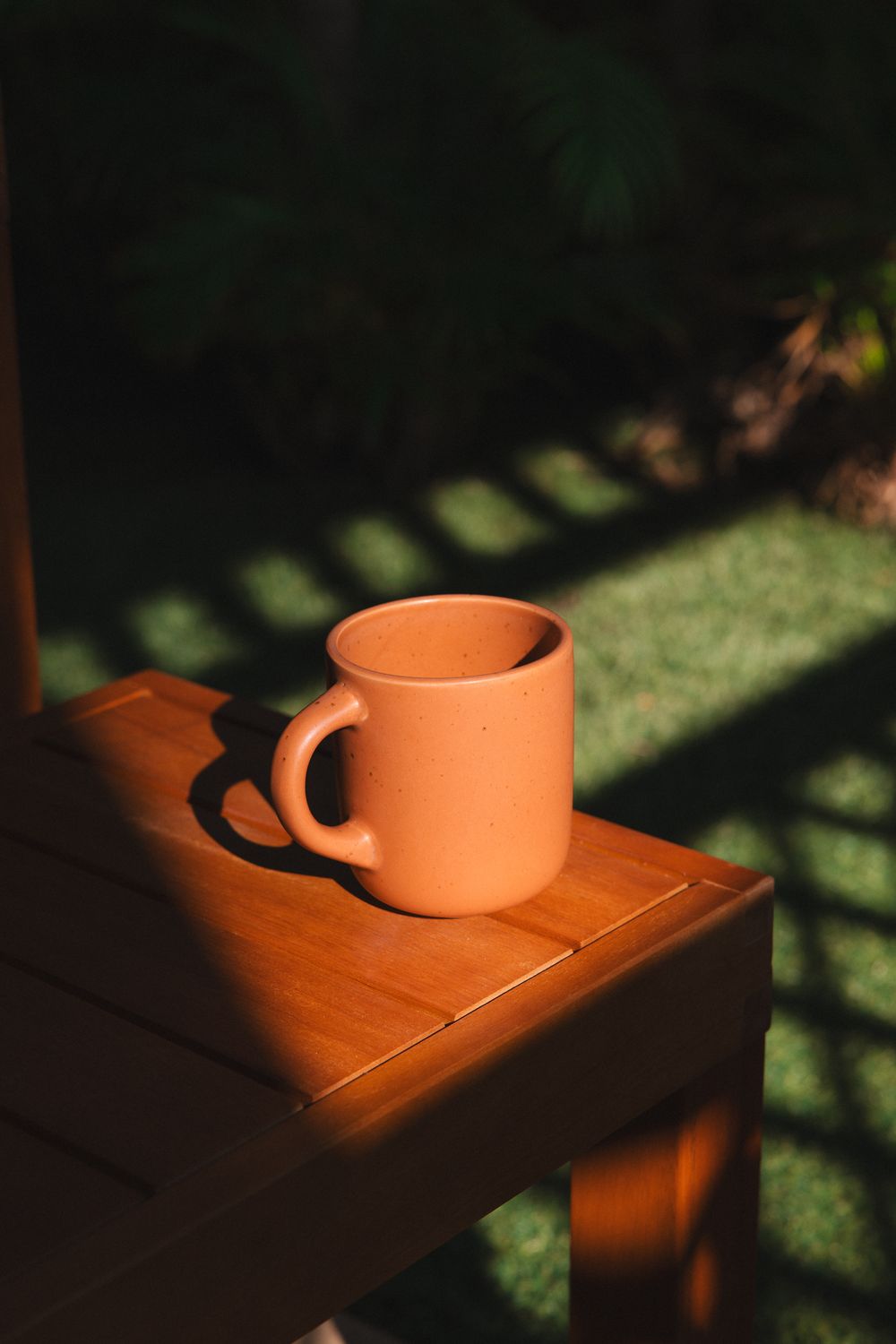 Terracotta clay mug sitting on a wooden chair arm in warm golden sunlight with garden background.