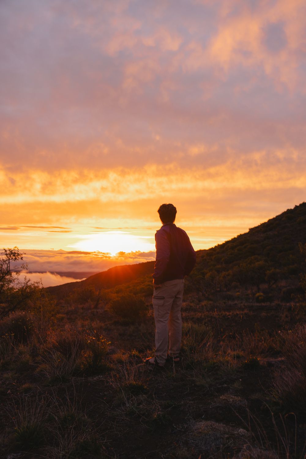 Silhouette of a person standing on a hillside watching a dramatic orange and purple mountain sunset.