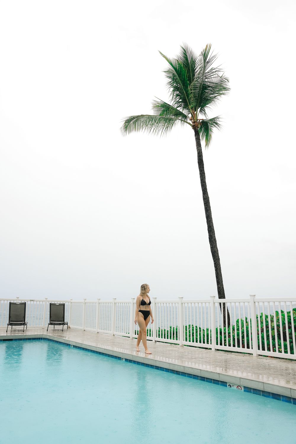 Woman in black bikini standing by a poolside with a tall palm tree and ocean view on an overcast day.