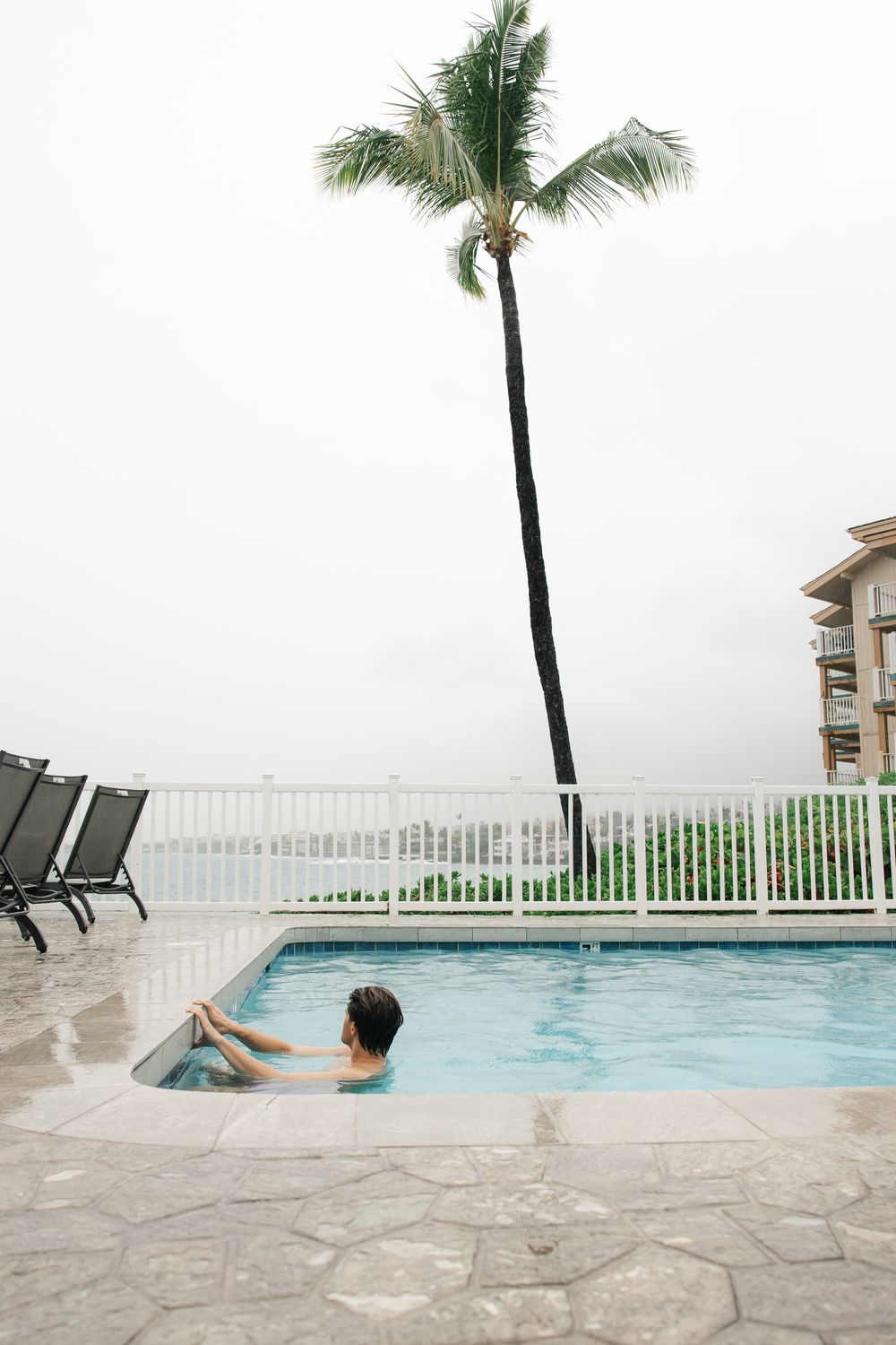 Person relaxing in a resort swimming pool with a tall palm tree and ocean view on an overcast day.