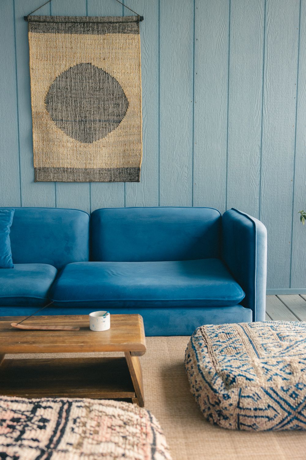 Cozy living room with blue velvet sofa, wooden coffee table, woven wall art, and bohemian floor cushion.