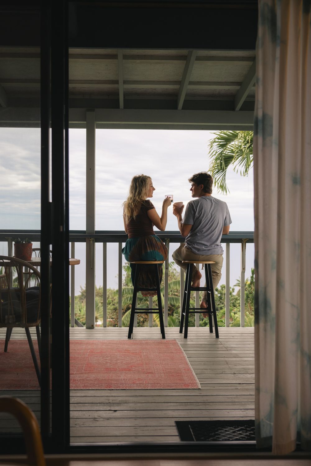 Couple sitting on bar stools on a tropical deck, enjoying drinks with a scenic ocean and palm tree view.