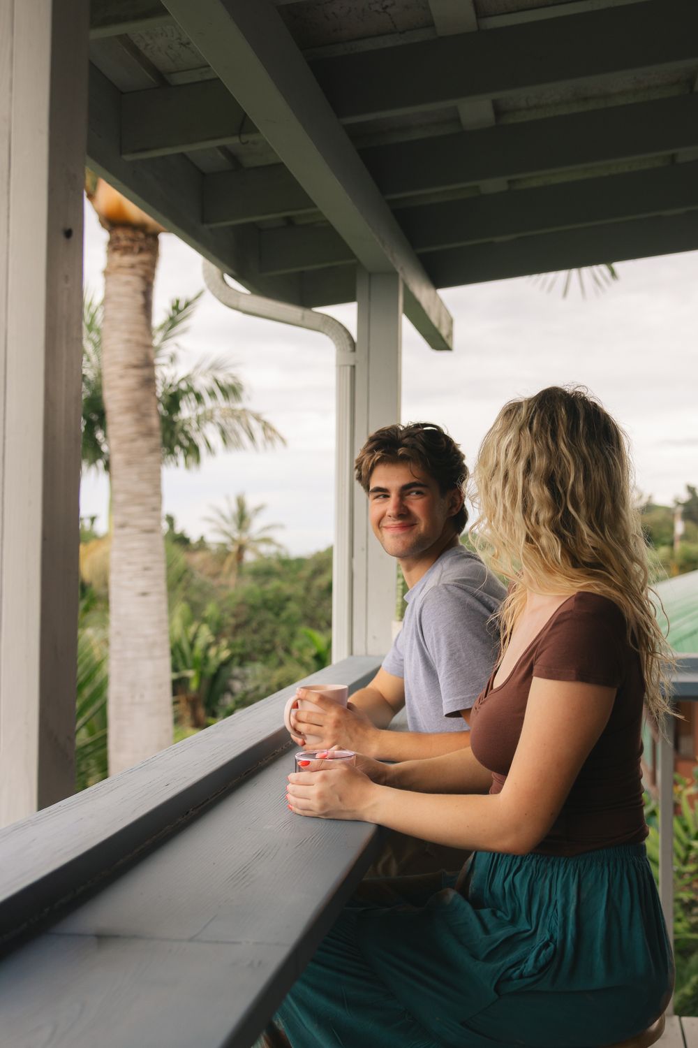 Smiling couple relaxing on a tropical porch balcony overlooking lush palm trees and greenery.