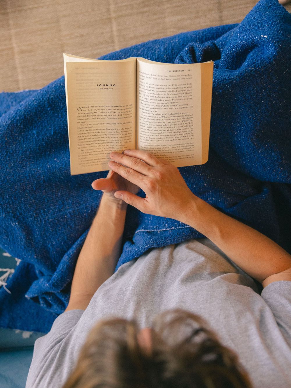 Person reading an open book while lying under a blue blanket, overhead view.