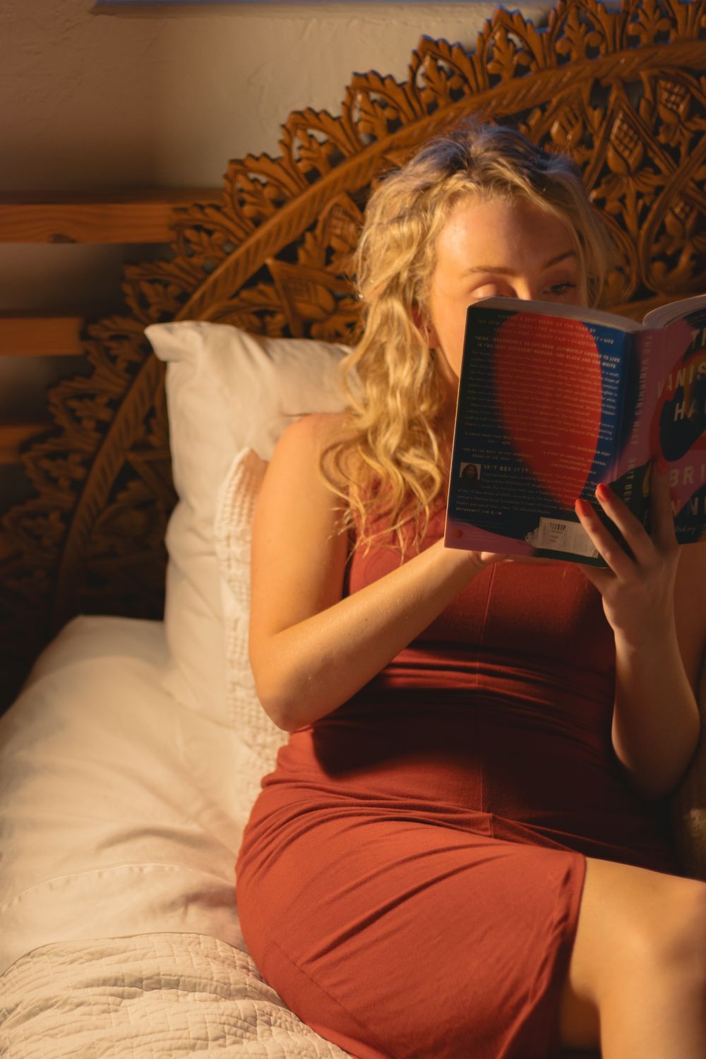 Blonde woman in red dress reading a book on a bed with ornate wooden headboard in warm golden light.