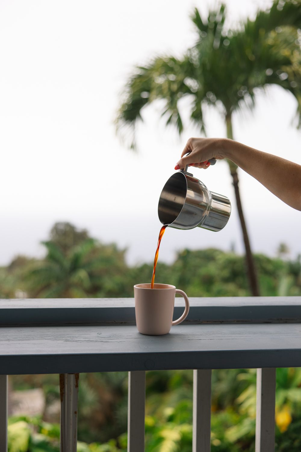 Hand pouring hot coffee from a steel pitcher into a pink mug on a balcony with tropical palm trees.
