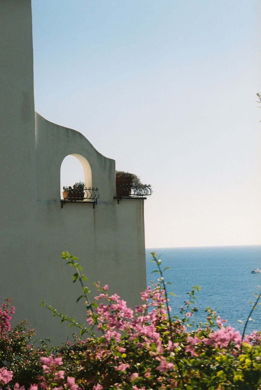 White Mediterranean building with arched window and balcony overlooking blue sea, framed by pink bougainvillea flowers.