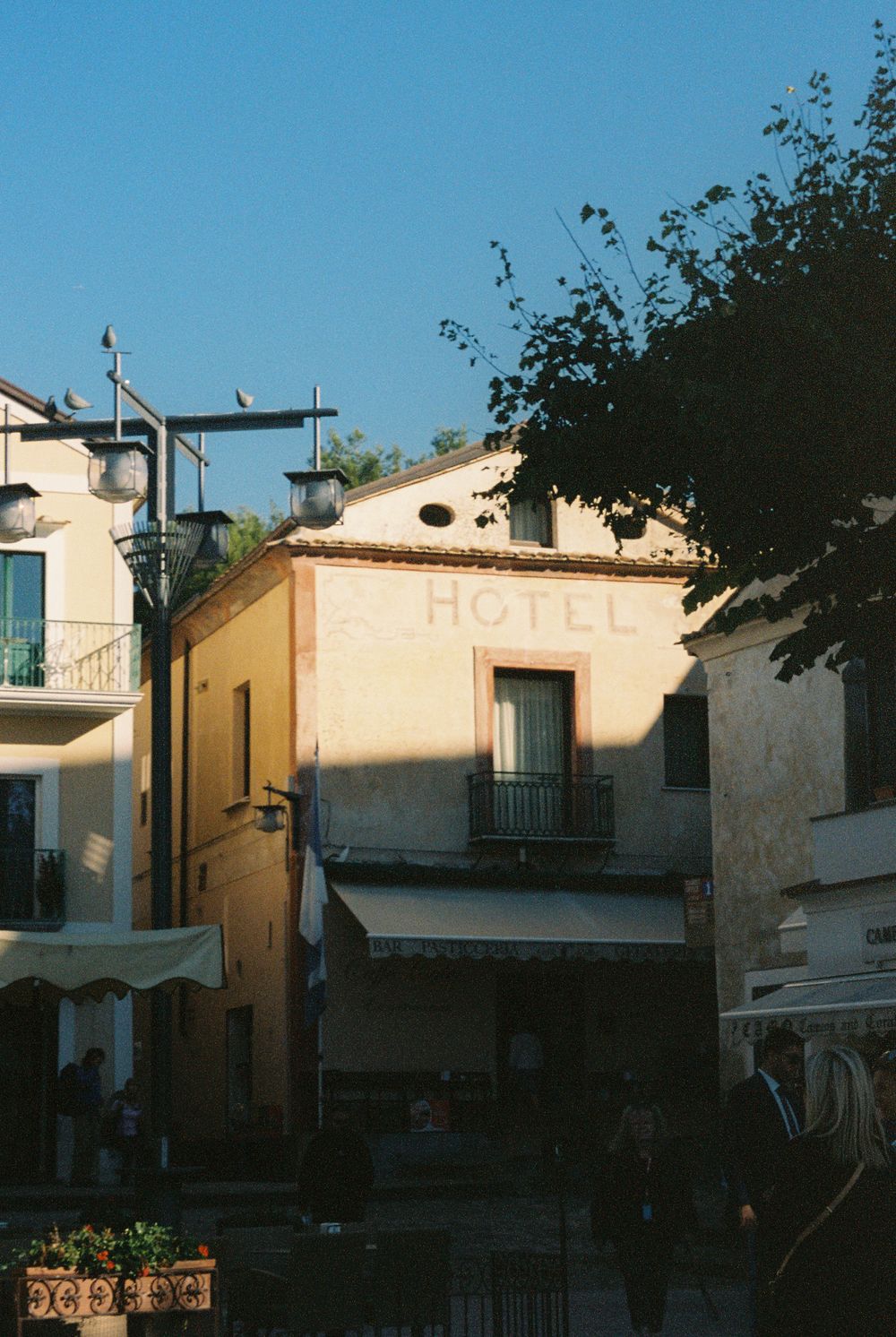 Charming Italian hotel building with yellow facade, balcony, street lamps, and lush trees under a clear blue sky.